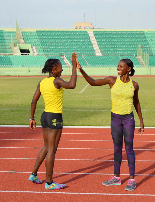 Two girls in athletic clothes give a high five as they stand on the track in a stadium.