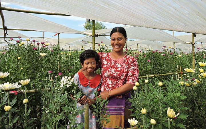 A woman and girl stand under a roof made of white fabric. They are surrounded by flowers.