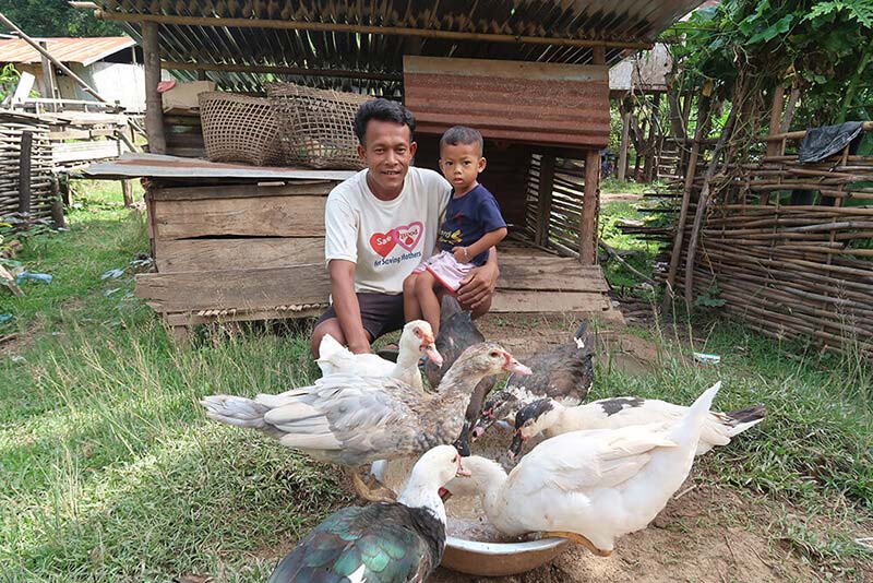 A man and his toddler smile posing in front of their chickens and ducks.