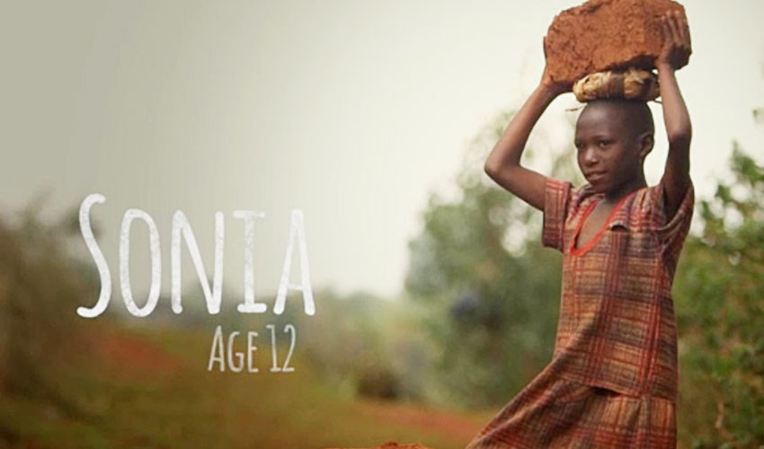 A young 12-year-old girl stands and carries a heavy stone on her head.