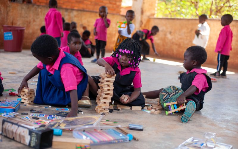 Three Malawian children sit together in a classroom, playing with Jenga blocks.