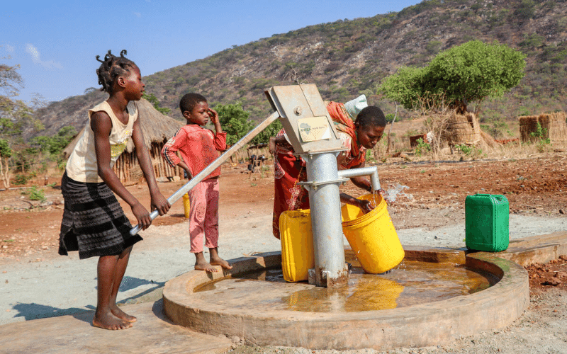 A girl pumps water into a yellow bucket held by a woman. A younger boy looks on.