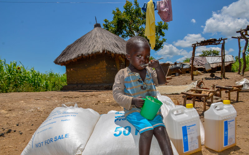 A child sits on top of sacks of food assistance eating some food. She is outside and there is a hut in the background and laundry hangs overhead.
