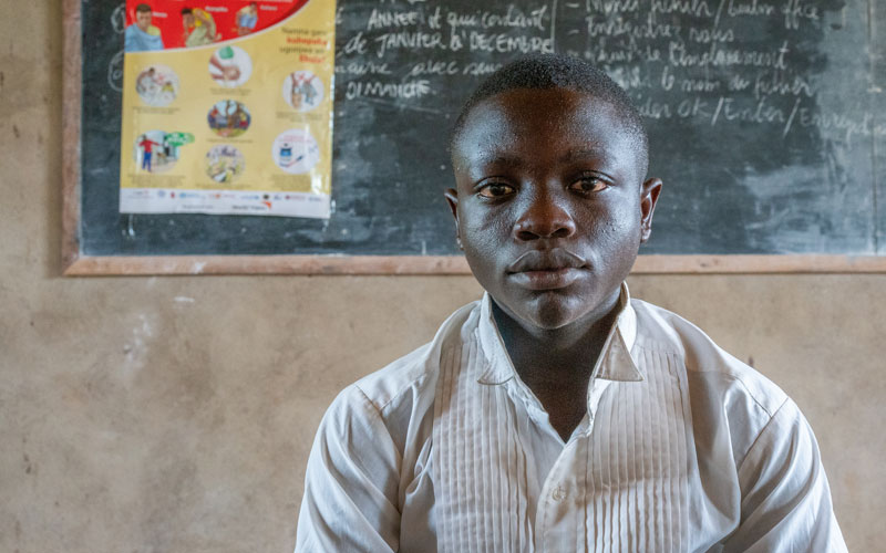 A boy from DRC sits in front of chalkboard with writing on it.