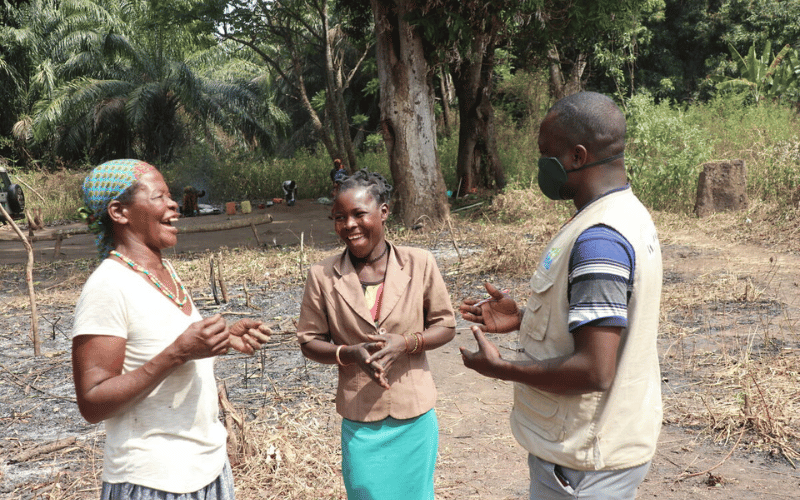 Mikelina and Severina speak and laugh with a World Vision staff member outside surrounded by trees.