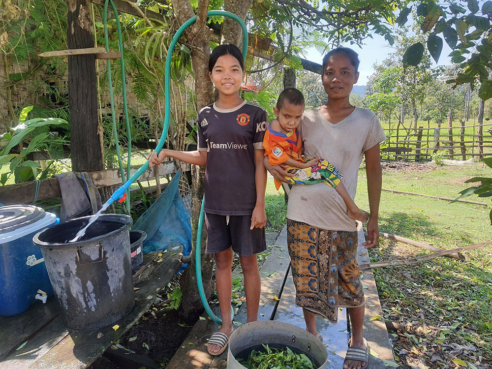 A young girl is smiling as she stands holding a hose that is filling a bucket with water. Beside her stands her mother holding a young boy. 