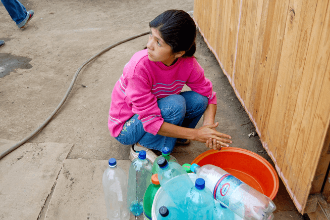 A child crouches to wash her hands in a bowl. Empty bottles of water stand beside her. 