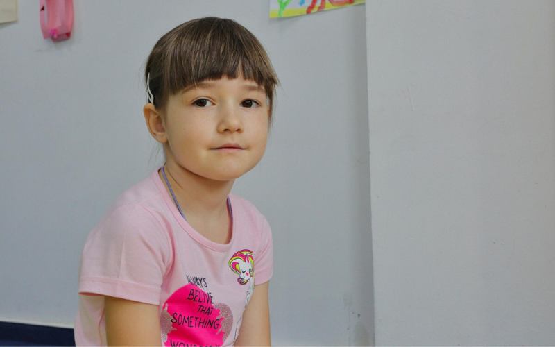 A very young girl wearing a pink shirt looks into the camera.