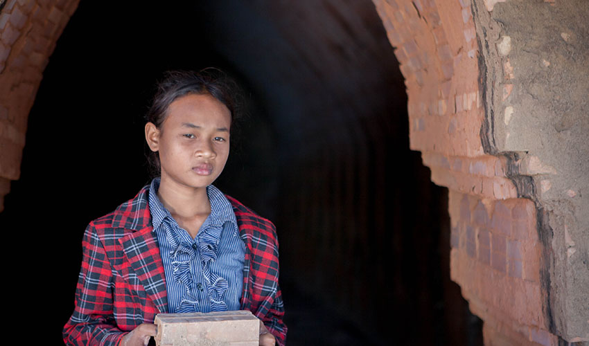 A cambodian girl holds a couple of clay bricks and stands in a doorway looking somber