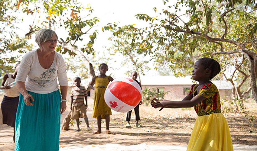 A Canadian woman and Tanzanian girl throw a beach ball with a maple leaf on it back and forth in a village.