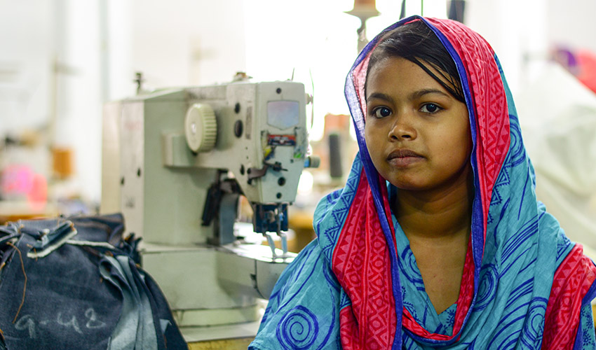 A young girl in Banlgadesh sits in a factory with a sewing machine behind her.