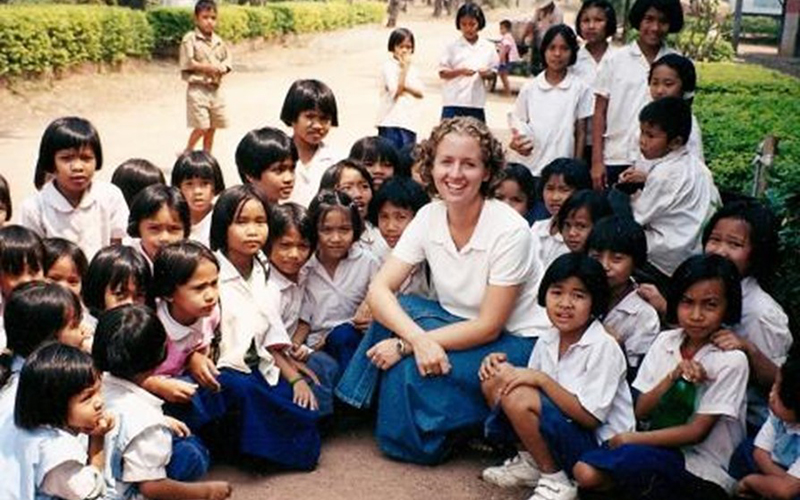 A woman sits amongst a group of young children in school uniforms.