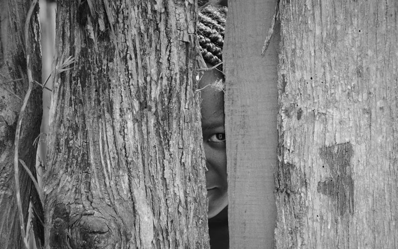 a young black girl looks through a wood fence. Only a sliver of her face around her eye is visible.