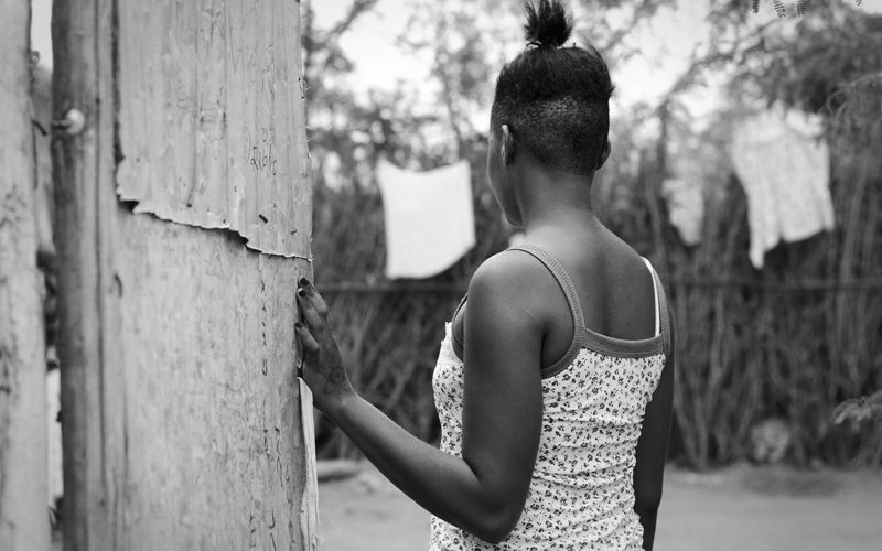 A young black girl stands with her back to the camera. She's looking into a fenced in yard, her left hand is on the gate.