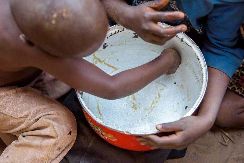 A little boy scrapes the last smears of food from inside a large bowl that’s held by an older person.