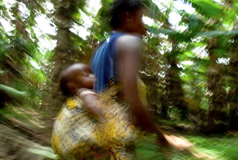 A blurry photo of a teen girl carrying her baby on her back in a yellow patterned sling, as she hurries through the jungle.