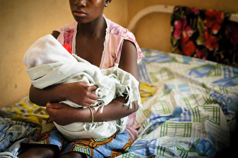 An adolescent girl holds a bundled baby in her arms, while sitting on a hospital bed. We don’t see the faces of either.