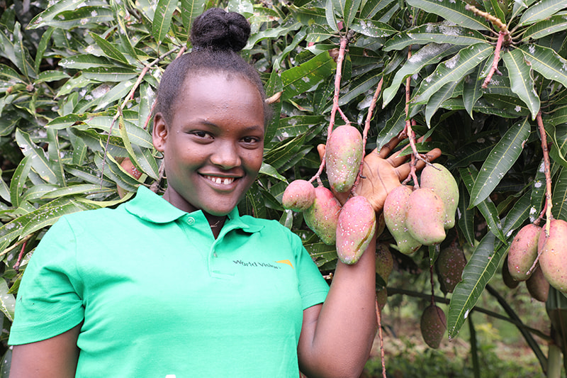 Une jeune femme portant un t-shirt vert montre fièrement des mangues poussant sur un manguier.