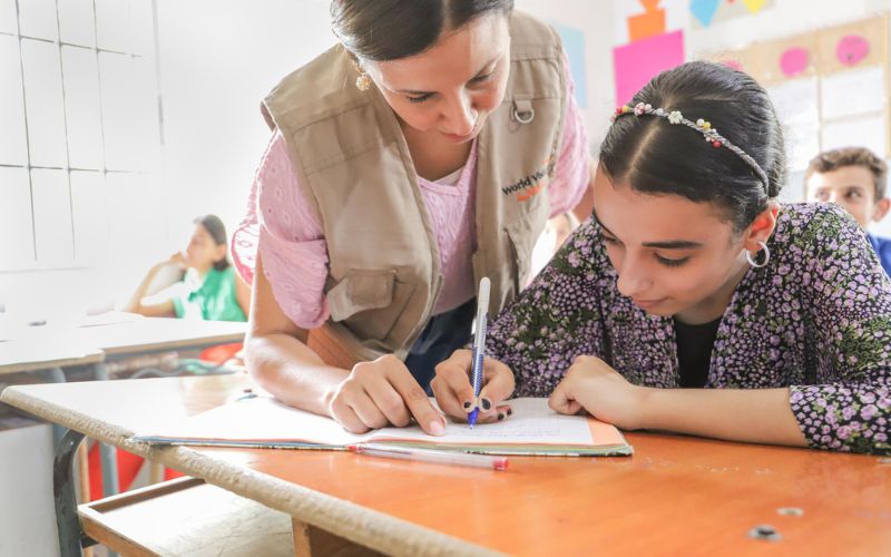 A woman wearing a World Vision vest leans over to help a girl who is writing on paper.