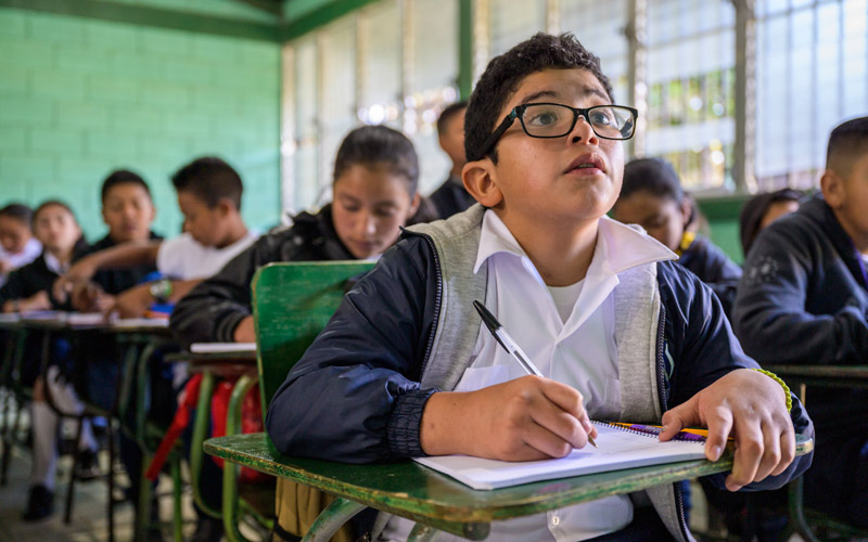 a young boy in Honduras looks up from his notebook at the blackboard. He is in a classroom.