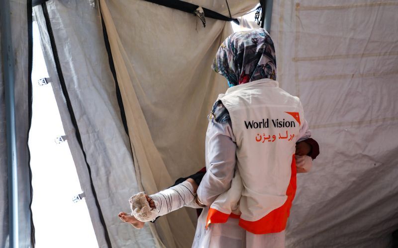 A World Vision worker carries a child with a splinted and bandaged leg out of a tent.