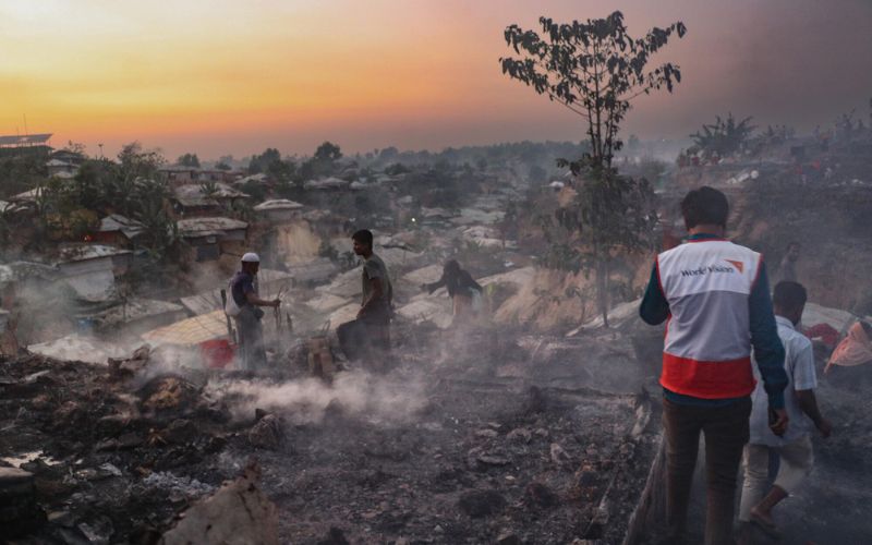 A man in a World Vision best walks toward the buildings of a refugee camp that are smoldering after a fire.