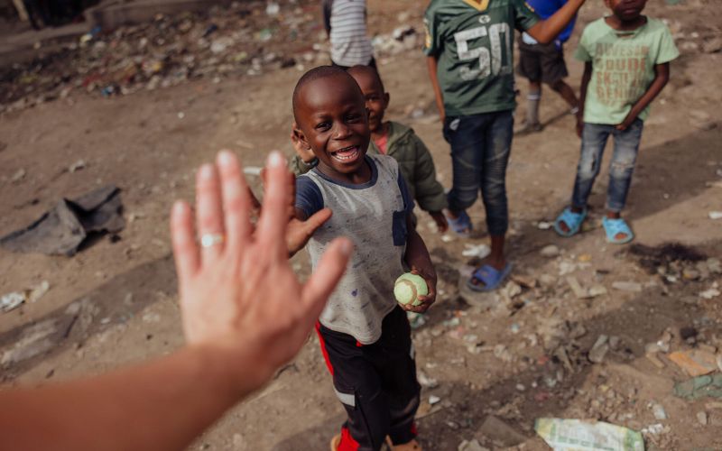 An adult high-fives with a small boy standing with his friends amidst rubble. The boy is holding a baseball and smiling.