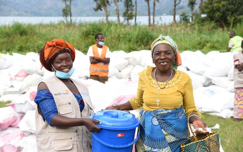 A woman in a World Vision best hands a pail of supplies to a woman. Both are smiling.