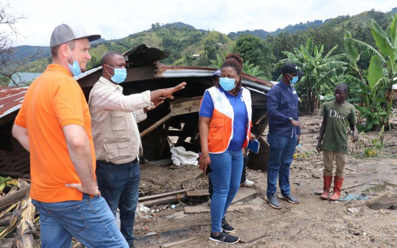 A man gestures to other adults to show  destruction from a natural disaster in a tropical region.
