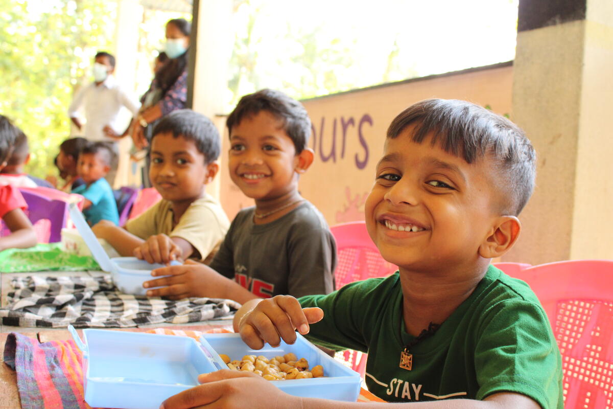 Children smile while eating lunch from blue containers at a long table.
