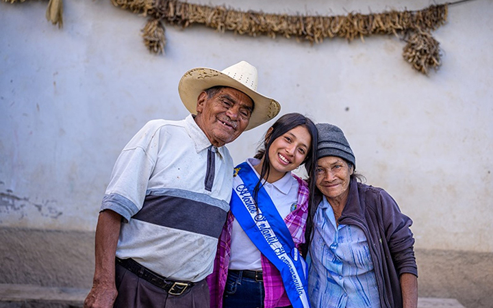 A young woman stands between an elderly man and an elderly woman. The three have their arms around one another and are smiling.