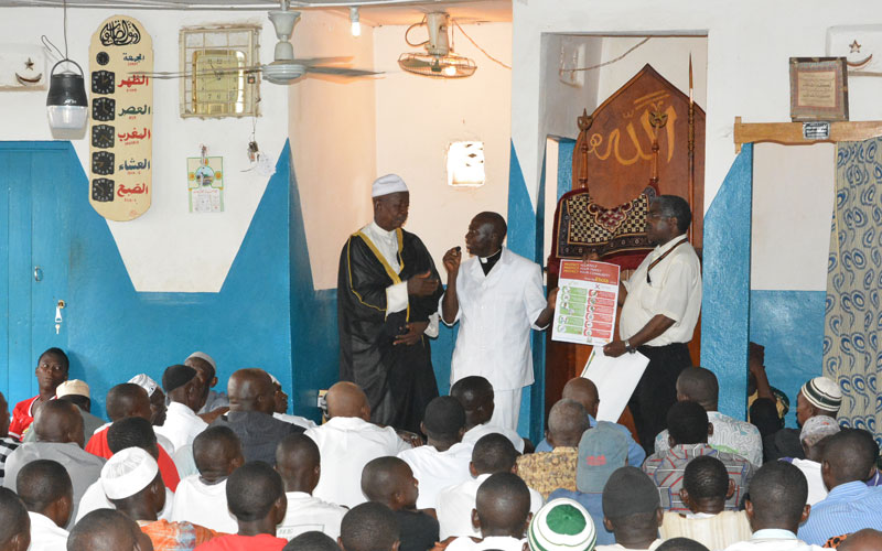 three men from Sierra Leone stand at the front of a mosque full of men.
