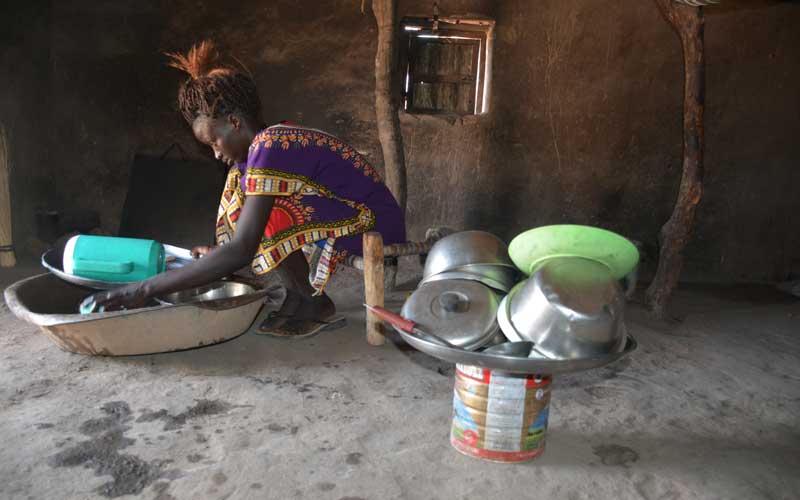 A young woman crouches over a large metal bowl, washing dishes. She’s in a hut with a dirt floor.