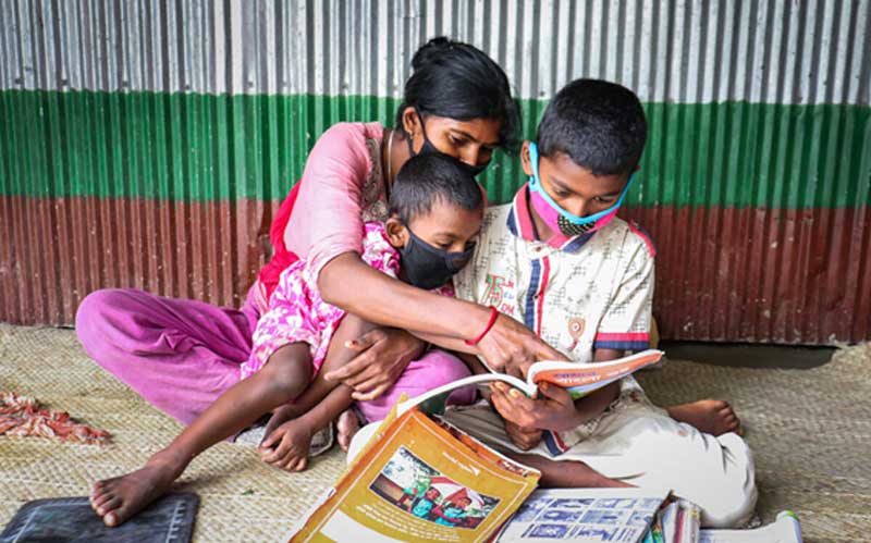 A mother and her two children, all wearing protective masks, read books together. They are sitting on the floor of their home.
