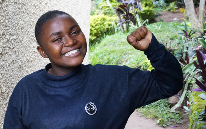 A young Congolese girl smiles at the camera. She is wearing a navy blue sweatshirt and is pumping her fist in the air jubilantly.