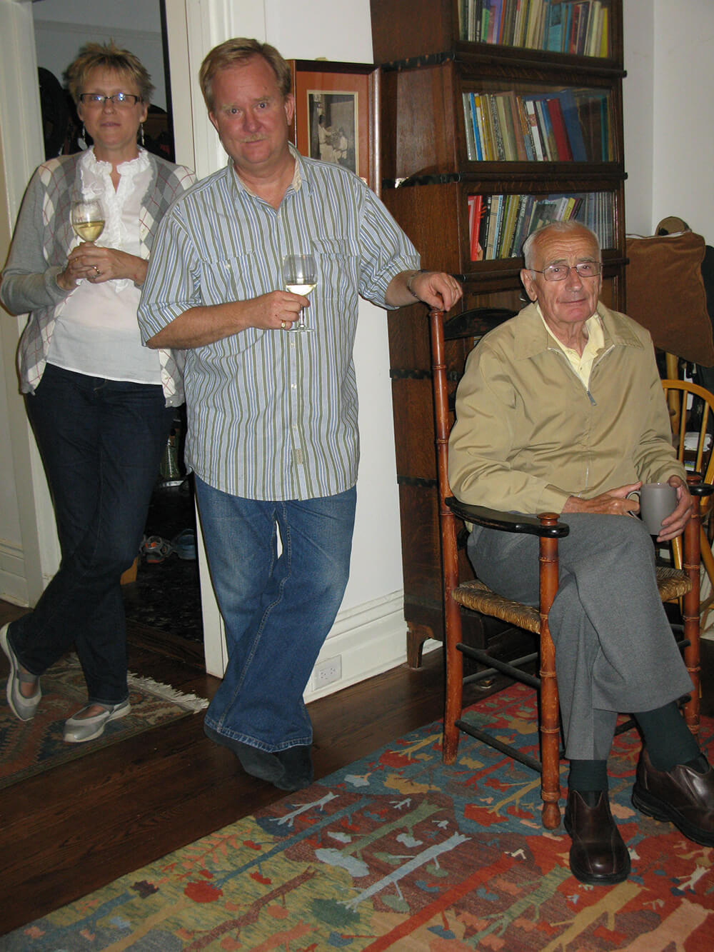 In this June 2012 photograph, we see Sandra, Tom, and Arie gathered in a study room. Arie is seated on a chair positioned in the bottom left of the picture. Standing in the middle is Tom, with his left arm casually resting on the chair that Arie occupies, while holding a wine glass in his right hand. Positioned behind Tom is Sandra, also holding a wine glass.