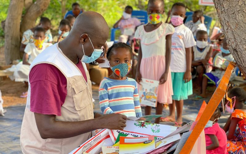 A man in a World Vision vest wears a protective mask while a line of children, wearing colourful masks and holding learning materials, look on.