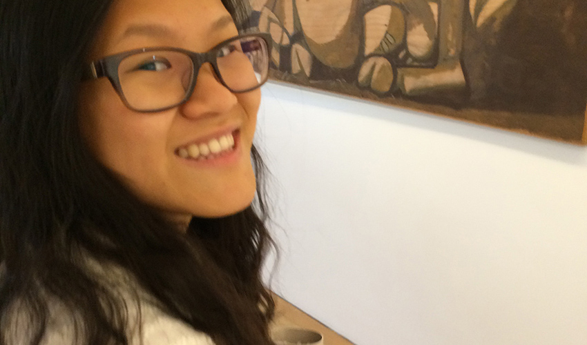 Young woman wearing black framed glasses next to a brown-toned art exhibit.
