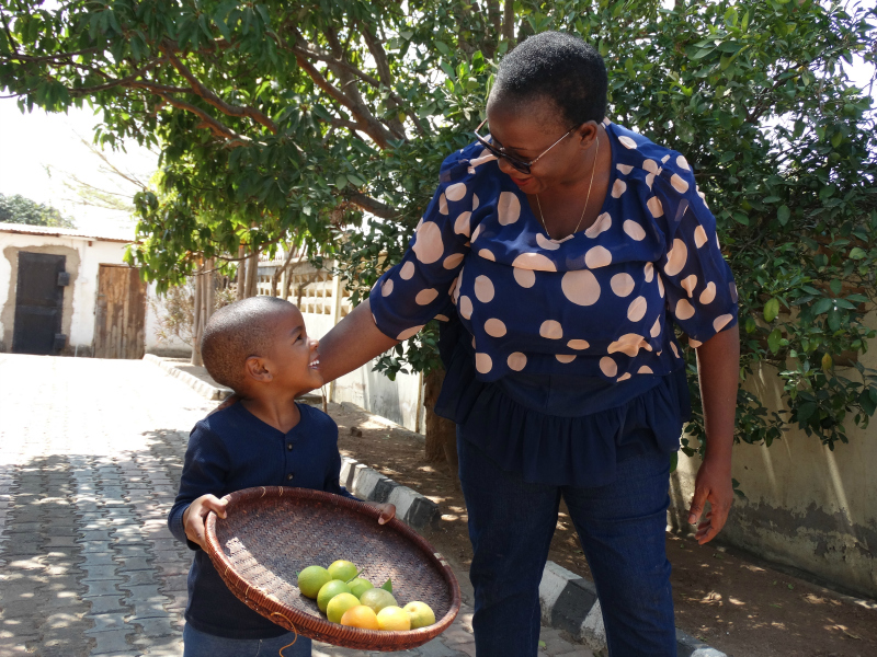 A child holds a basket of fruit and smiles up at her mother.