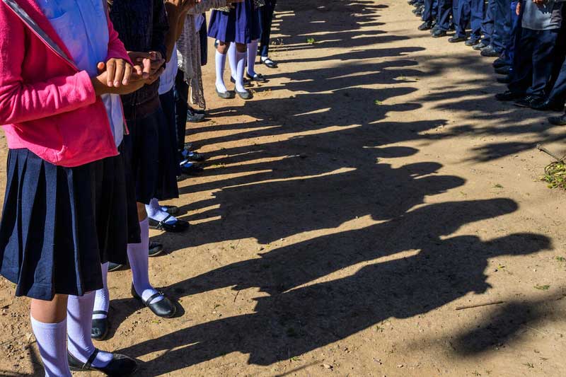 A row of shadows is cast on the dirt ground, cast by a row of girls in school uniforms. The photo does not show the children’s faces.