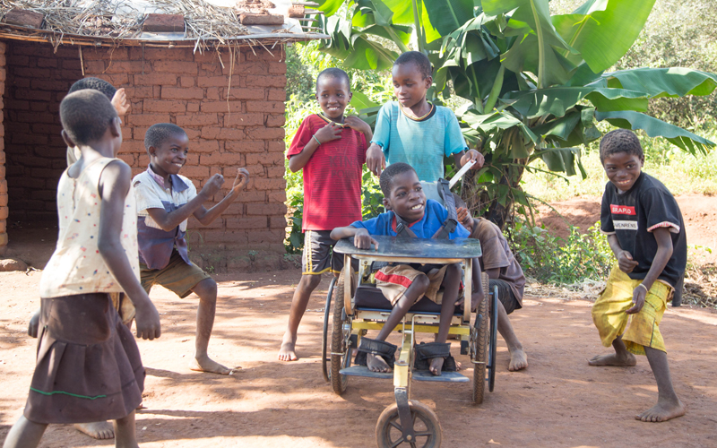 A group of children from Malawi play in front of a child's home. One of the children is in a wheelchair.