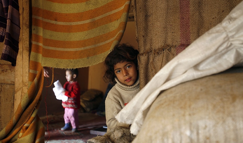 A little girl living in a Syrian refugee camp, peeks from behind worn and tattered tapestries..
