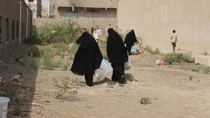 Three women wearing niqabs walk across a barren space between buildings, carrying bags of hygiene kits.