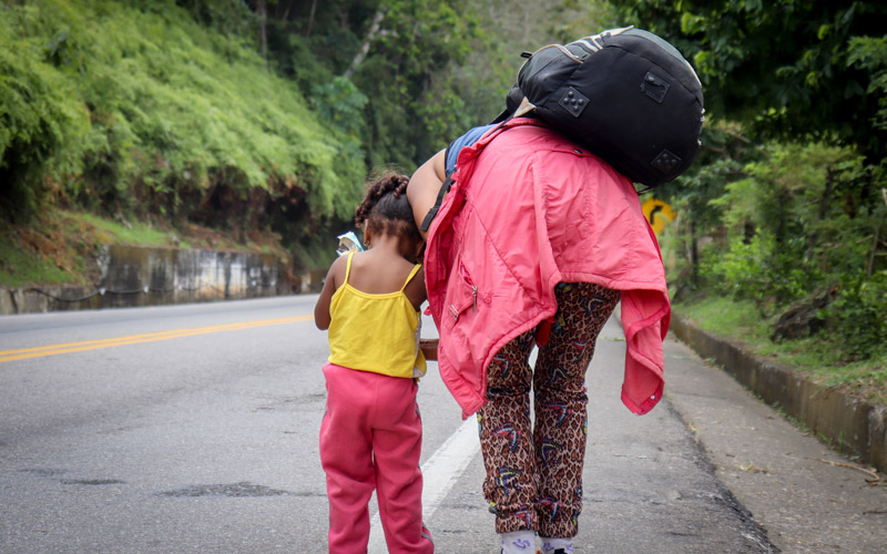A Venezuelan woman crouches down to the little girl beside her. They are at the side of a green mountain road.