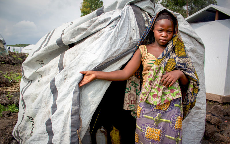 A young Black girl holds open the entrance to the tent she shares with friends in a refugee camp in DRC.