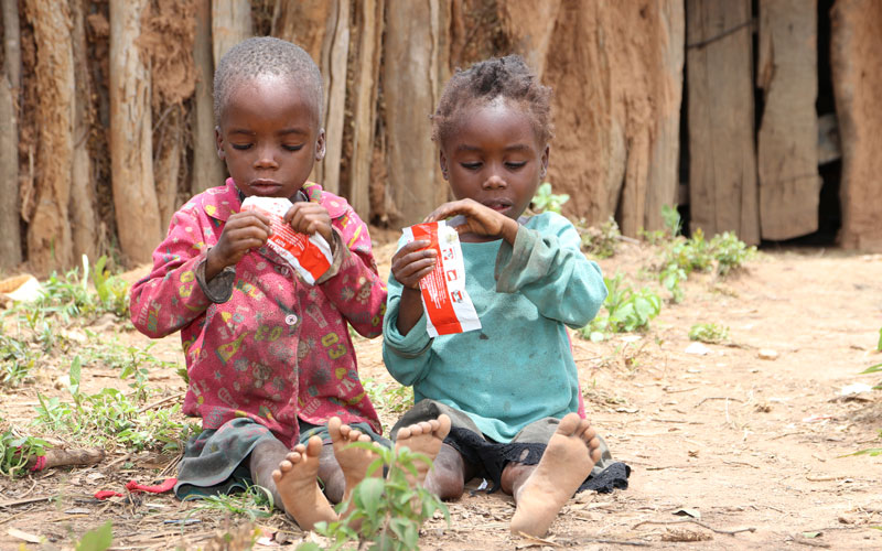 A girl and a boy sitting on the ground, eating one pack each of ready-to-use therapeutic food.