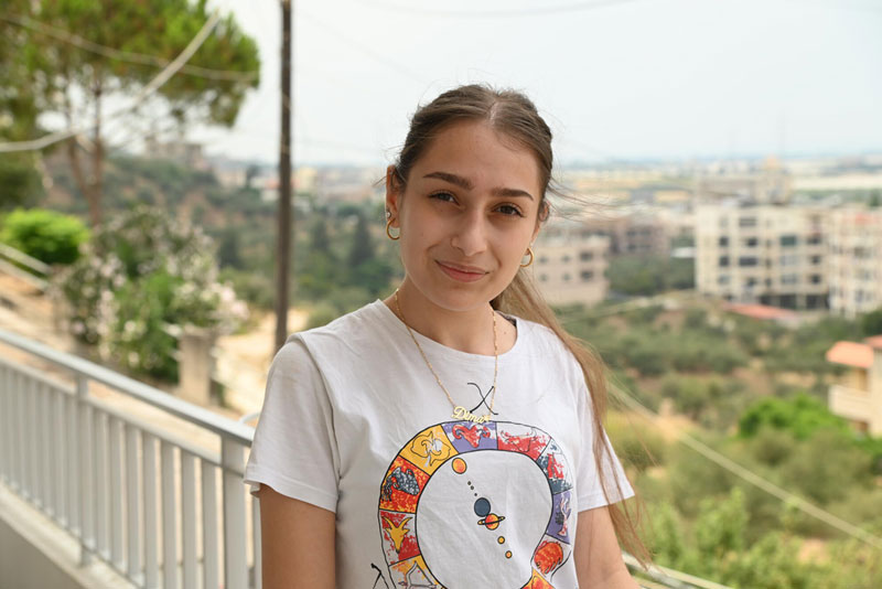 A teenage girl standing in front of a balcony railing wearing a white graphic t-shirt and smiling for a picture.