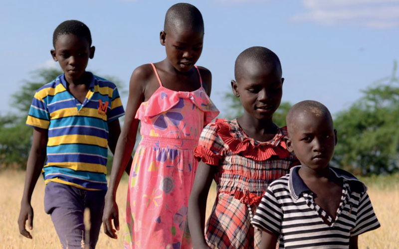 Two African girls and two boys walk in a line through a field of tall, dried-out grass.