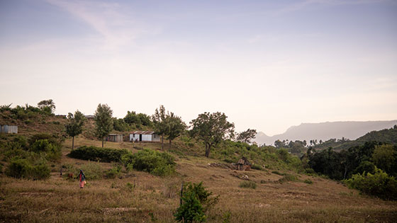 A woman walking on mountainous land surrounded by sprawling trees and few houses.