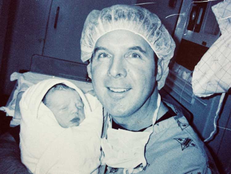 A man wearing surgical clothing carrying a newborn baby.